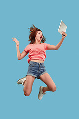 Image showing Image of young woman over blue background using laptop computer or tablet gadget while jumping.