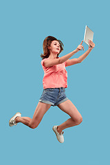 Image showing Image of young woman over blue background using laptop computer or tablet gadget while jumping.