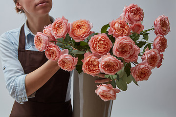 Image showing Girl florist in a brown apron with a vase of pink roses on a gray background.