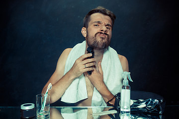Image showing The young man in bedroom sitting in front of the mirror scratching his beard