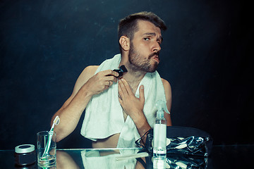 Image showing The young man in bedroom sitting in front of the mirror scratching his beard