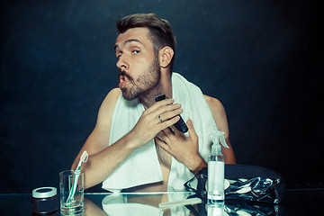 Image showing The young man in bedroom sitting in front of the mirror scratching his beard