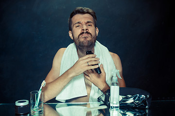 Image showing The young man in bedroom sitting in front of the mirror scratching his beard