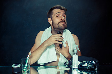 Image showing The young man in bedroom sitting in front of the mirror scratching his beard