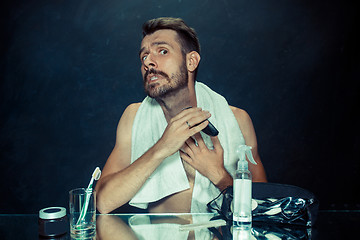 Image showing The young man in bedroom sitting in front of the mirror scratching his beard