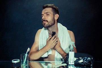 Image showing The young man in bedroom sitting in front of the mirror scratching his beard