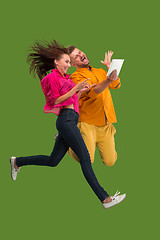 Image showing Jump of young couple over green studio background using laptop or tablet gadget while jumping.