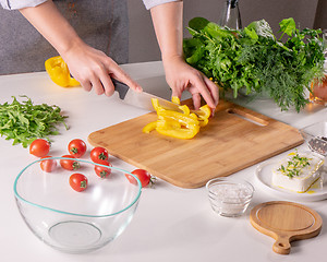 Image showing A woman\'s hand slices a yellow pepper on a wooden board on a white kitchen table. Preparation of salad