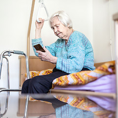 Image showing Elderly 96 years old woman reading phone message while sitting on medical bed supporting her by holder.