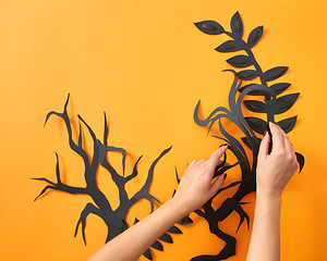 Image showing The hands of a woman make a composition of paper black leaves and trees on an orange background with space for text. Handdecraft lyout to Halloween. Flat lay