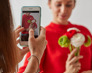 Image showing The girl takes a photo on the phone of a young pretty woman with a bouquet of fresh flowers on a gray background.