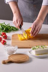 Image showing The girl\'s hand cuts pieces of pepper on a wooden board, cheese, and tomatoes on a white kitchen table. Preparation of salad