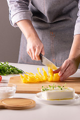Image showing Pieces of pepper on a wooden board, female hands cut vegetable for salad on a white kitchen table