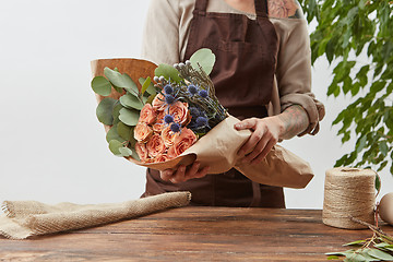 Image showing Creating of beautiful flowers bouquet from living coral color roses with green leaves in a paper in a woman\'s hands with tattoo on a gray background.