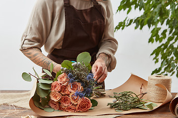 Image showing Florist at workspace. Woman\'s hands with tattoo are making bouquet from fresh natural flowers coral living color with decorative leaves on a wooden table step by step.