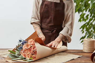 Image showing Florist woman is wrapping fresh rose bouquet in a decorative paper on a wooden table on a light wall background. Process step by step. Mother\'s Day holiday.