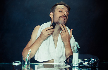 Image showing young man in bedroom sitting in front of the mirror scratching his beard