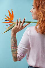 Image showing Girl with a tattoo holding on her shoulder an orange flower strelitzia around a blue background with space for text. Creative layout