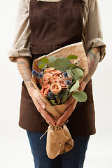 Image showing Woman in a brown apron is holding summer wonderful fresh roses, eryngium and green leaves as a greeting bunch on a light background. Greeting card. Copy space.