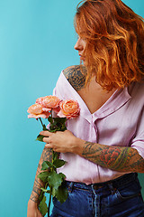Image showing Pretty woman with red hair and tattoo on her shoulders, hands in a pink shirt holds bouquet from fresh roses on a blue background. Copy space.