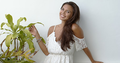 Image showing Happy brunette in white dress standing next to green plant