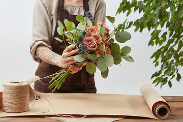 Image showing Young woman florist is making bouquet with fresh flowers roses living coral color at the table with paper and rope. Process step by step. Concept floral shop.