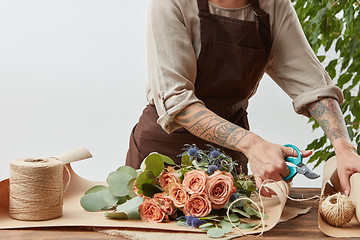 Image showing Female florist is cutting paper for creating beautiful bouquet on a background of light wall. Place for text. Process of making bouquet step by step.