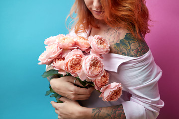 Image showing Attractive red-haired girl with tattoo on shoulders and hands holds a bunch of coral roses on a blue-pink background. A gift for Mother\'s Day.