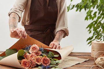 Image showing Gardener girl make bouquet for a holiday from living coral roses and branches of green leaf on a wooden table on a light background. Mother\'s Day.