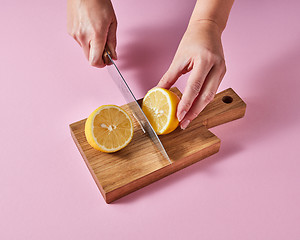 Image showing Woman\'s hands cut a lemon on a wooden board with a knife around a pink background with copy space. Ingredient for summer drink