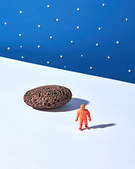 Image showing Plastic toy of spaceman and moonstone on a light surface against blue star sky background.