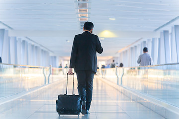 Image showing Businessman walking and wheeling a trolley suitcase at the lobby, talking on a mobile phone. Business travel concept.