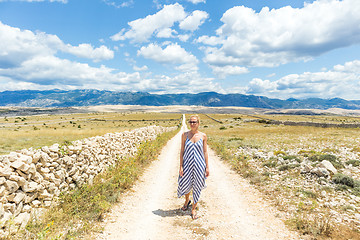 Image showing Caucasian young woman in summer dress holding bouquet of lavender flowers while walking outdoor through dry rocky Mediterranean Croatian coast lanscape on Pag island in summertime