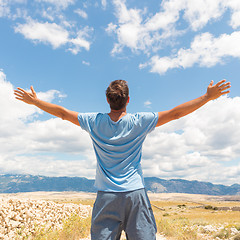 Image showing Rear view of casual sporty man standing on a dirt country road rising hands up to the clouds on a blue summer sky. Freedom and travel adventure concept.