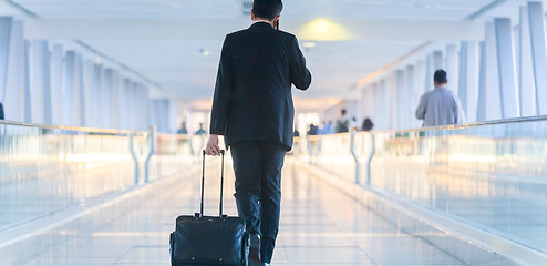 Image showing Businessman walking and wheeling a trolley suitcase at the lobby, talking on a mobile phone. Business travel concept.