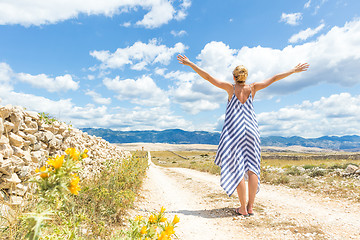 Image showing Rear view of woman in summer dress holding bouquet of lavender flowers while walking outdoor through dry rocky Mediterranean Croatian coast lanscape on Pag island in summertime