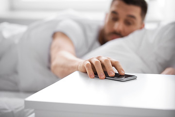 Image showing sleepy young man reaching for smartphone in bed