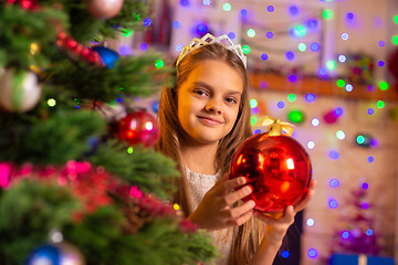 Image showing Beautiful ten year old girl peeking out with a big red ball from behind a Christmas tree