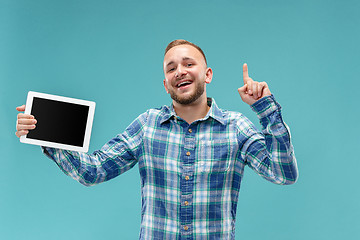 Image showing Studio picture of positive man isolated on blue background standing in casual clothes holding tablet and showing it blank screen with happy smile