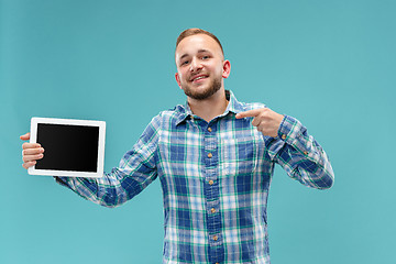 Image showing Studio picture of positive man isolated on blue background standing in casual clothes holding tablet and showing it blank screen with happy smile