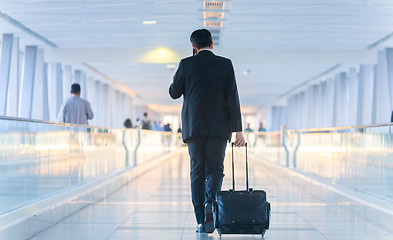 Image showing Businessman walking and wheeling a trolley suitcase at the lobby, talking on a mobile phone. Business travel concept.