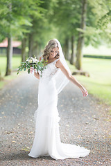 Image showing Full length portrait of beautiful sensual young blond bride in long white wedding dress and veil, holding bouquet outdoors in natural background