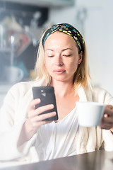 Image showing Beautiful caucasian woman at home, feeling comfortable wearing white bathrobe, taking some time to herself, drinking morning coffee and reading news on mobile phone device in the morning