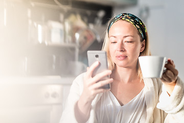 Image showing Beautiful caucasian woman at home, feeling comfortable wearing white bathrobe, taking some time to herself, drinking morning coffee and reading news on mobile phone device in the morning