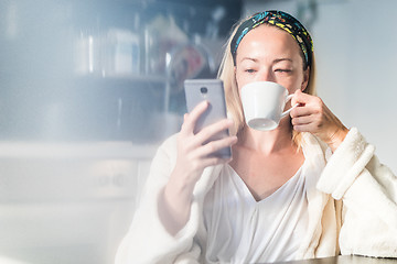 Image showing Beautiful caucasian woman at home, feeling comfortable wearing white bathrobe, taking some time to herself, drinking morning coffee and reading news on mobile phone device in the morning