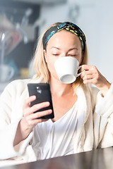 Image showing Beautiful caucasian woman at home, feeling comfortable wearing white bathrobe, taking some time to herself, drinking morning coffee and reading news on mobile phone device in the morning