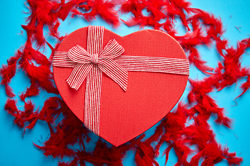 Image showing Red, heart shaped gift box placed on blue background among red feathers