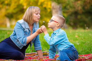 Image showing Mom and boy teen sit on green glade and blow off dandelion