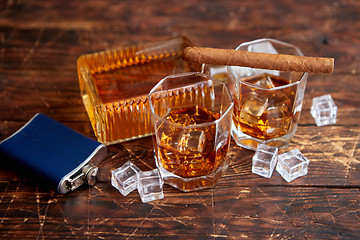 Image showing Bottle of whiskey with two glasses and cuban cigar placed on rustic wooden table