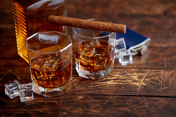 Image showing Bottle of whiskey with two glasses and cuban cigar placed on rustic wooden table